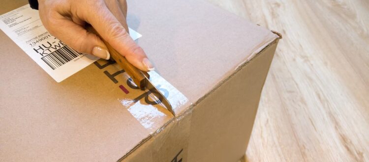 A person carefully cuts tape on a box using a knife. The image captures the motion of the person in action.